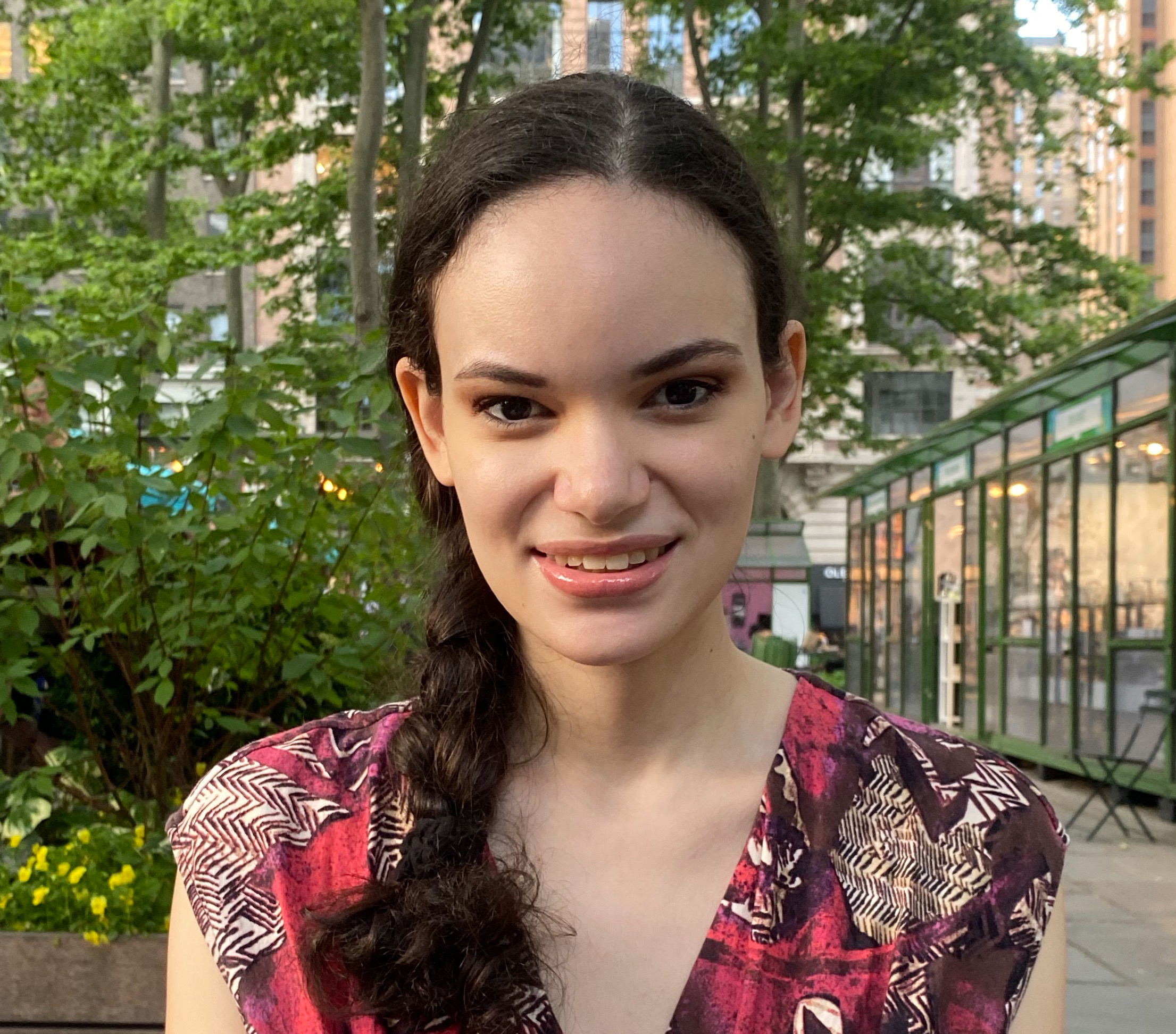 A picture of Zola Ray from the shoulders up, smiling in a red and brown patterned shirt. Her hair is in a braid and there is greenery in the background.